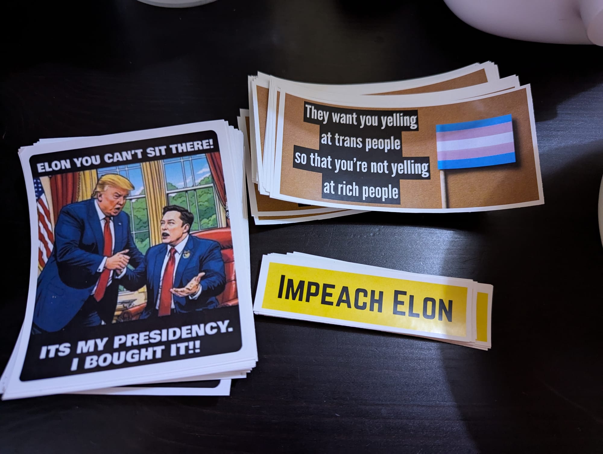 Three piles of stickers. One shows Elon Musk sitting at the Oval Office Desk while Trump tells him that he can't sit there. He replies "It's my presidency, I bought it!!" Another pile of stickers has a picture of the trans pride flag and text reading "They want you yelling at trans people so that you're not yelling at rich people". The third shows a yellow rectangle with black text reading "Impeach Elon".