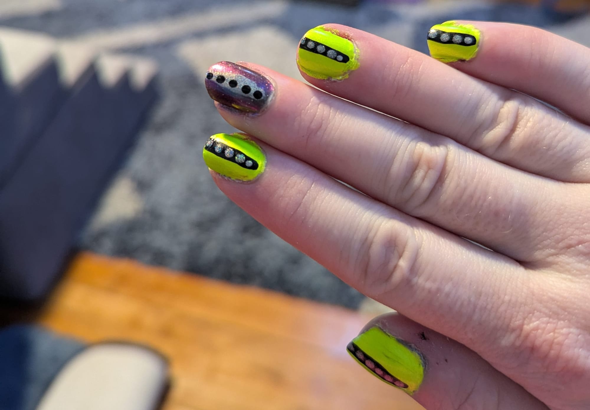 A picture of a light-skinned hand with elaborately painted nails. On most fingers, the background is a shimmery neon green, with a black stripe down the center and silver holographic dots along the stripe. On the middle finger, the background is a blue/purple duochrome with silver holo sparkles. This nail features a silver holo center stripe with black dots.
