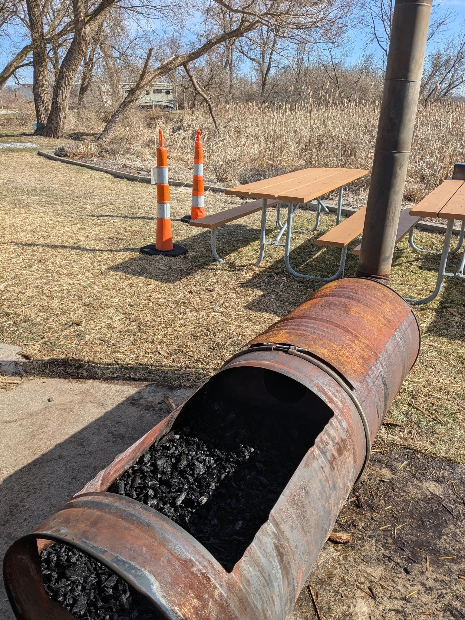 Two fifty-five gallon steel drums have been bolted together with a curved band of metal. The top of one has been cut out and uncovered, showing it is full of charcoal.