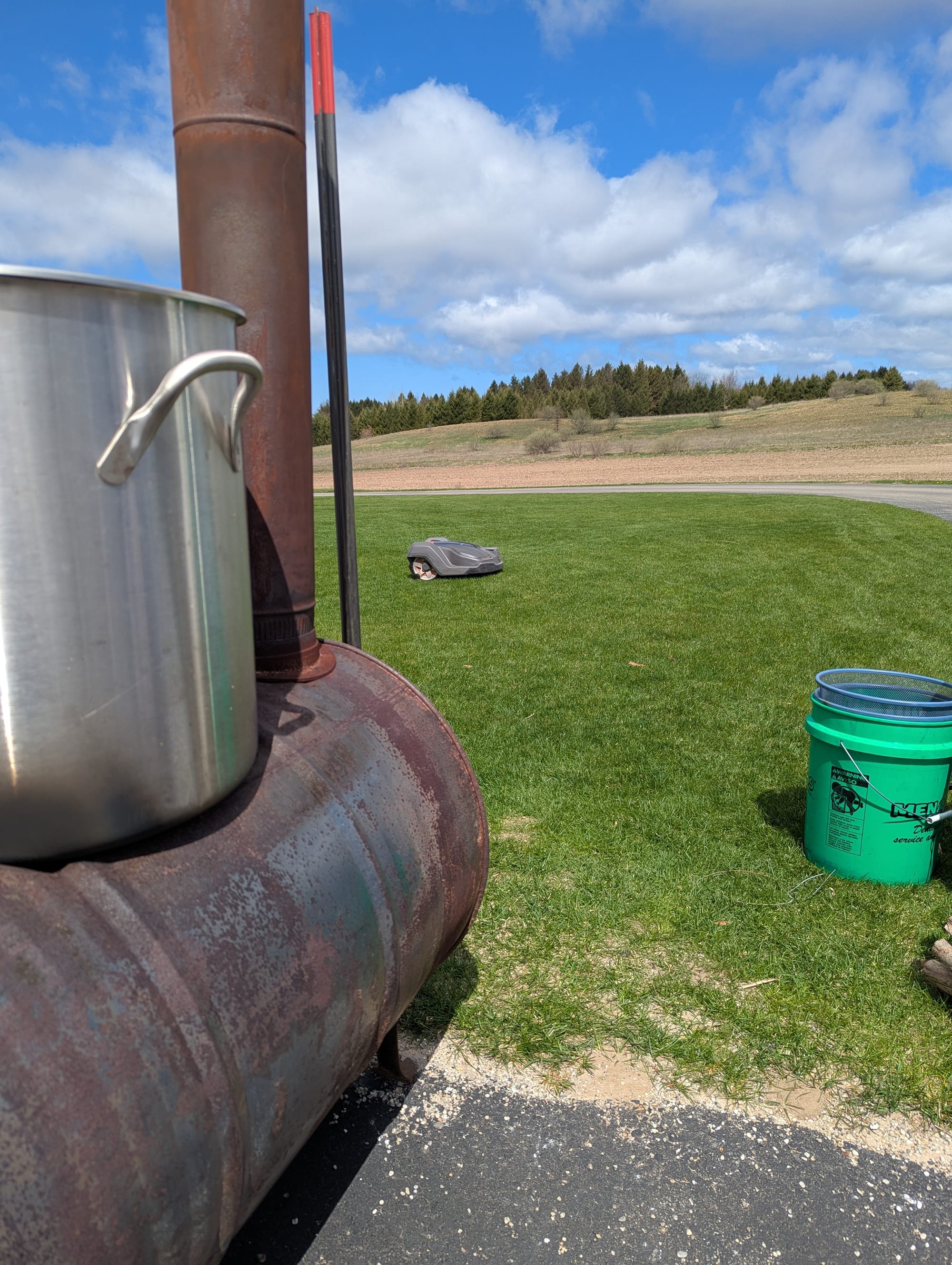 A biochar stove with a large stockpot on top of it sits at the edge of a parking lot. In the background, a robotic lawnmower that is clearly up to something attempts to sneak up on us.