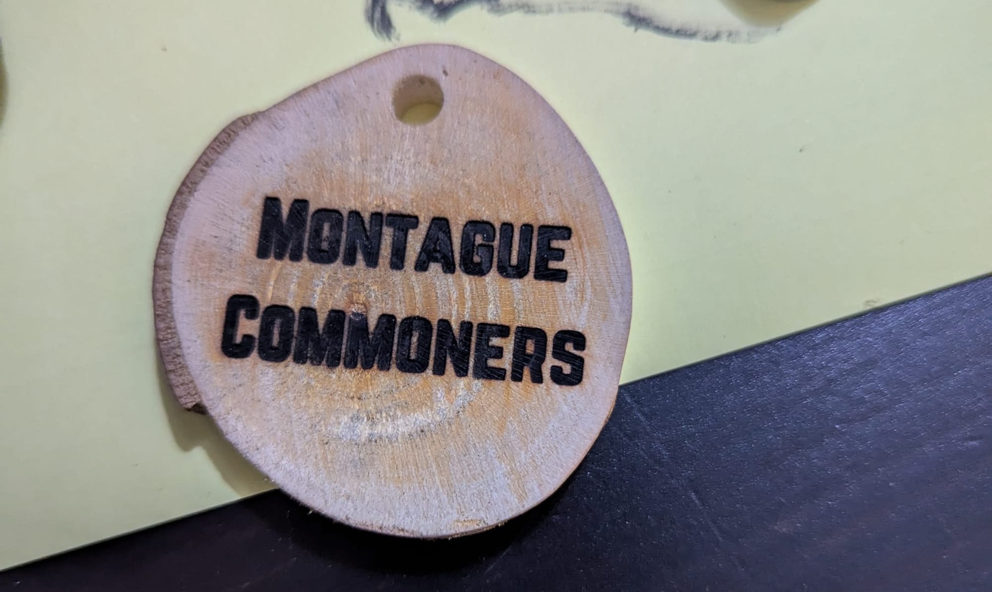 A small slice of log that has been sanded and stamped with a woodburning stamp. The stamp reads "Montague Commoners"