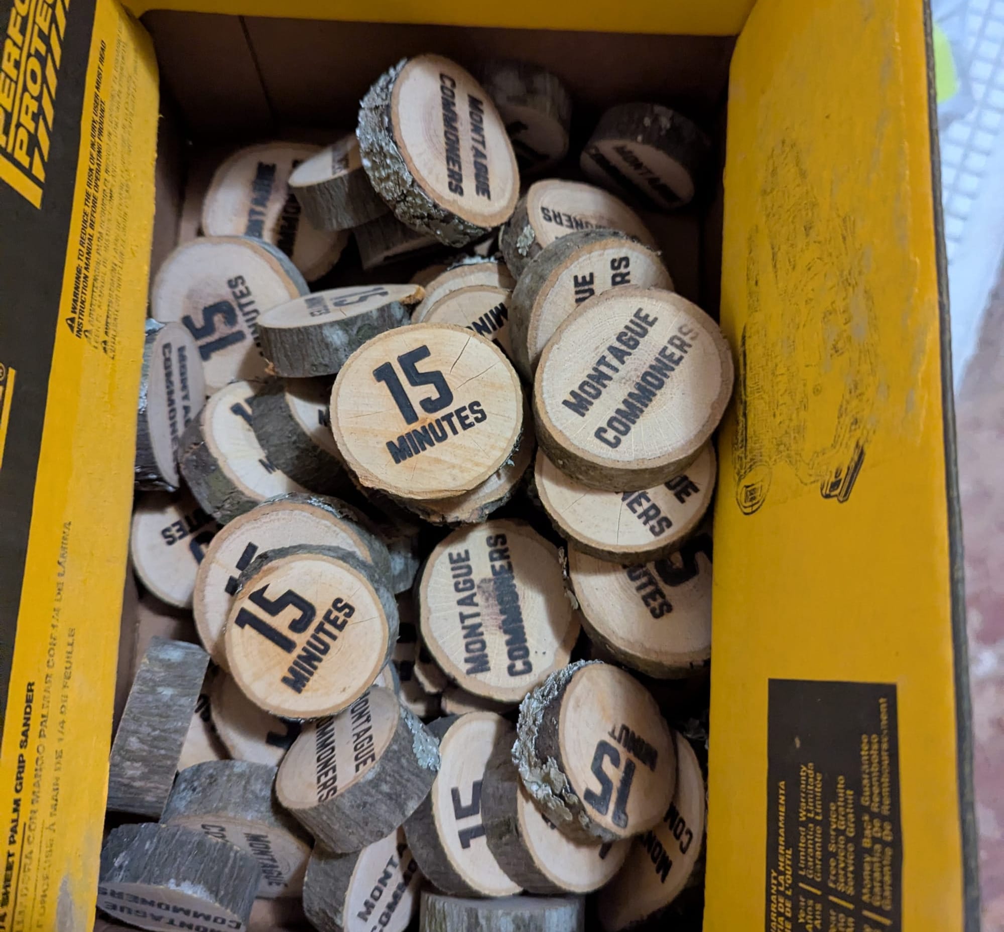 A box full of small slices of wood cut from a branch. They have lettering burned into them that says "Montague Commoners" on one side, and "15 minutes" on the other.