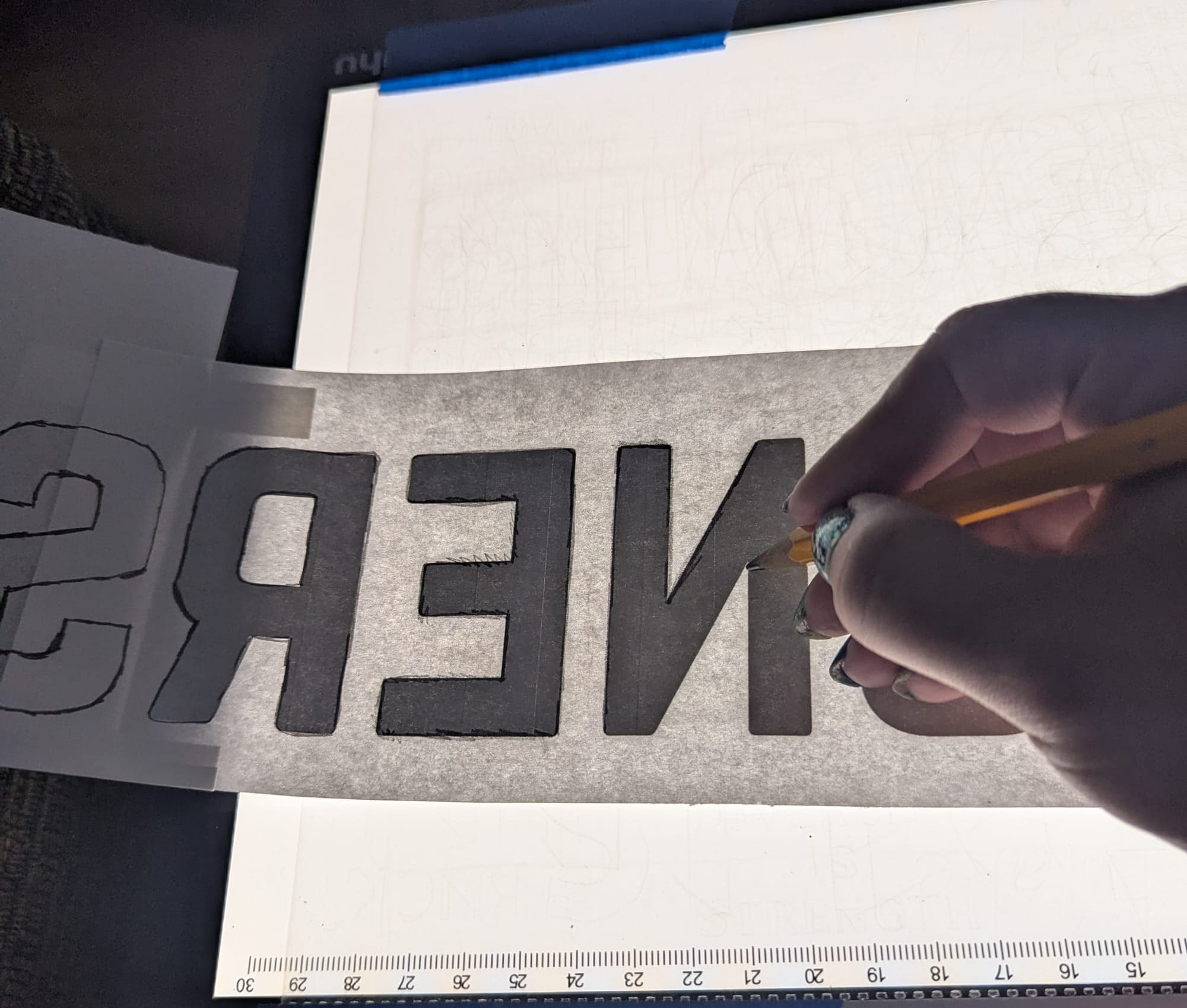 A piece of paper with the letters "n e r s" is upside down on a light pad. Wiley's hand is tracing the edges of the letters with a heavy pencil line in order to transfer the outlines to the sign.