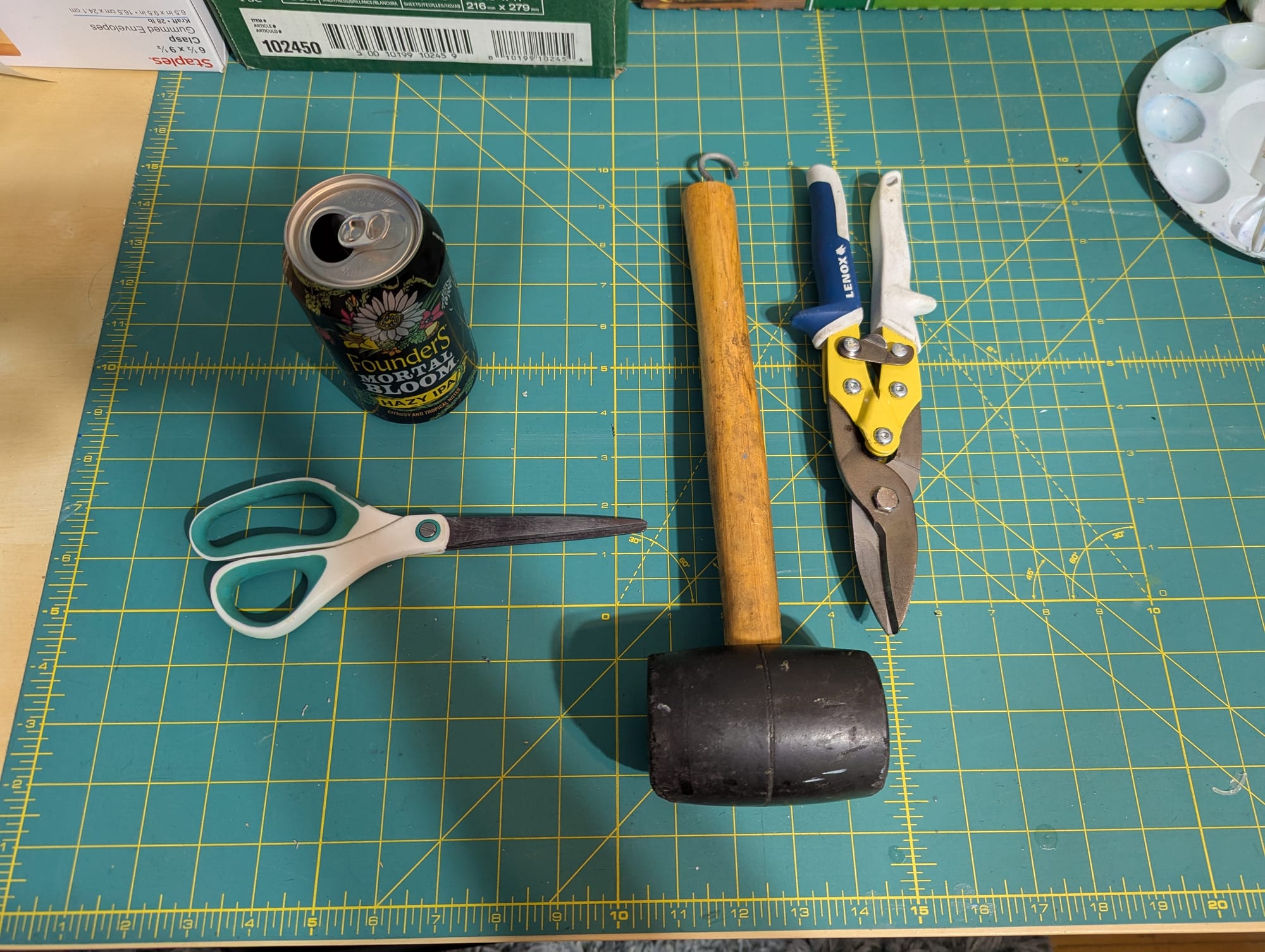An aluminum can, a pair of scissors, a rubber mallet, and a pair of tin snips arranged on a work mat