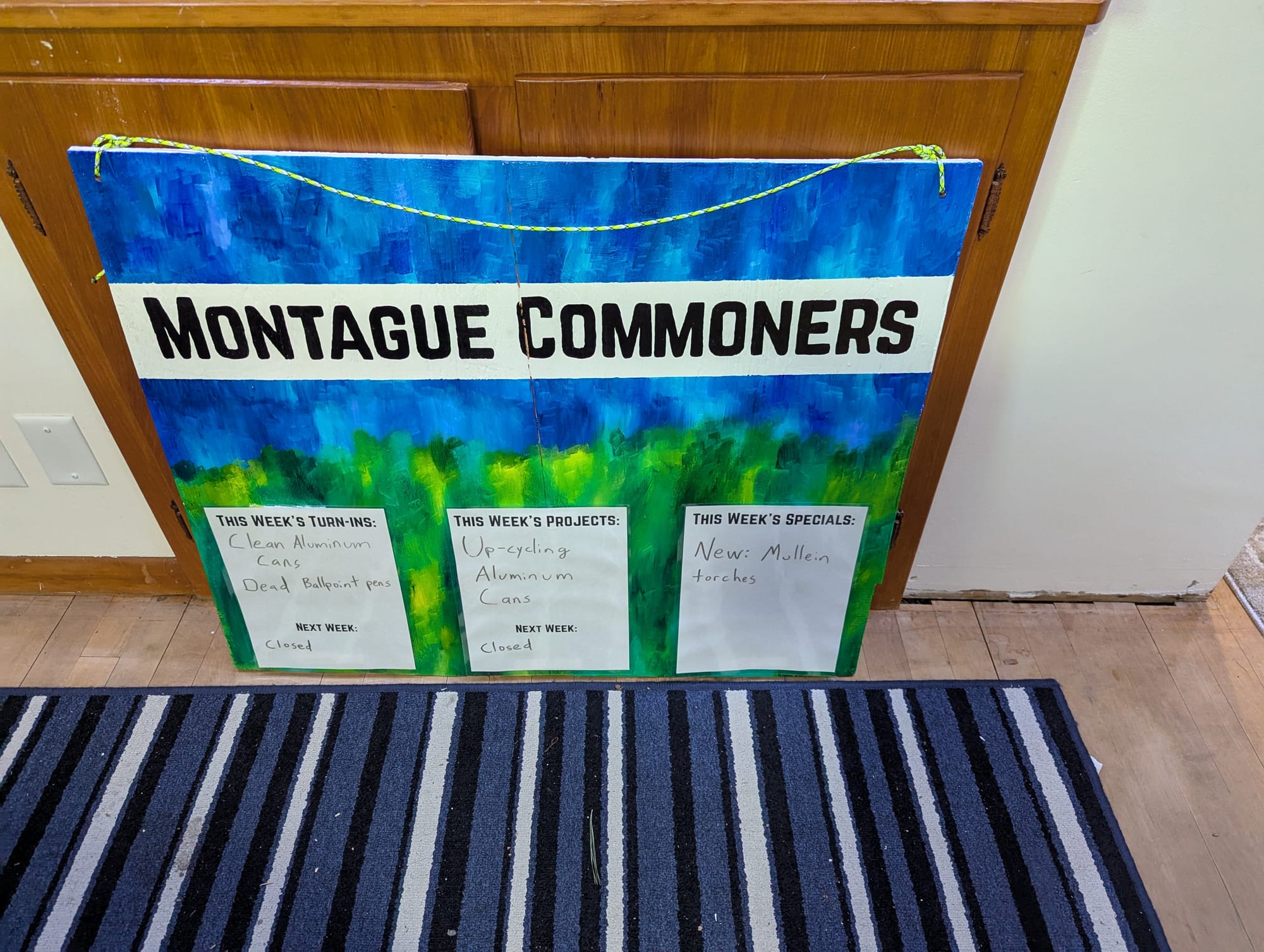 The Montague Commoners sign from the Artisan Market, now with the background painted in variegated blues and greens to resemble a blue sky over green plant life