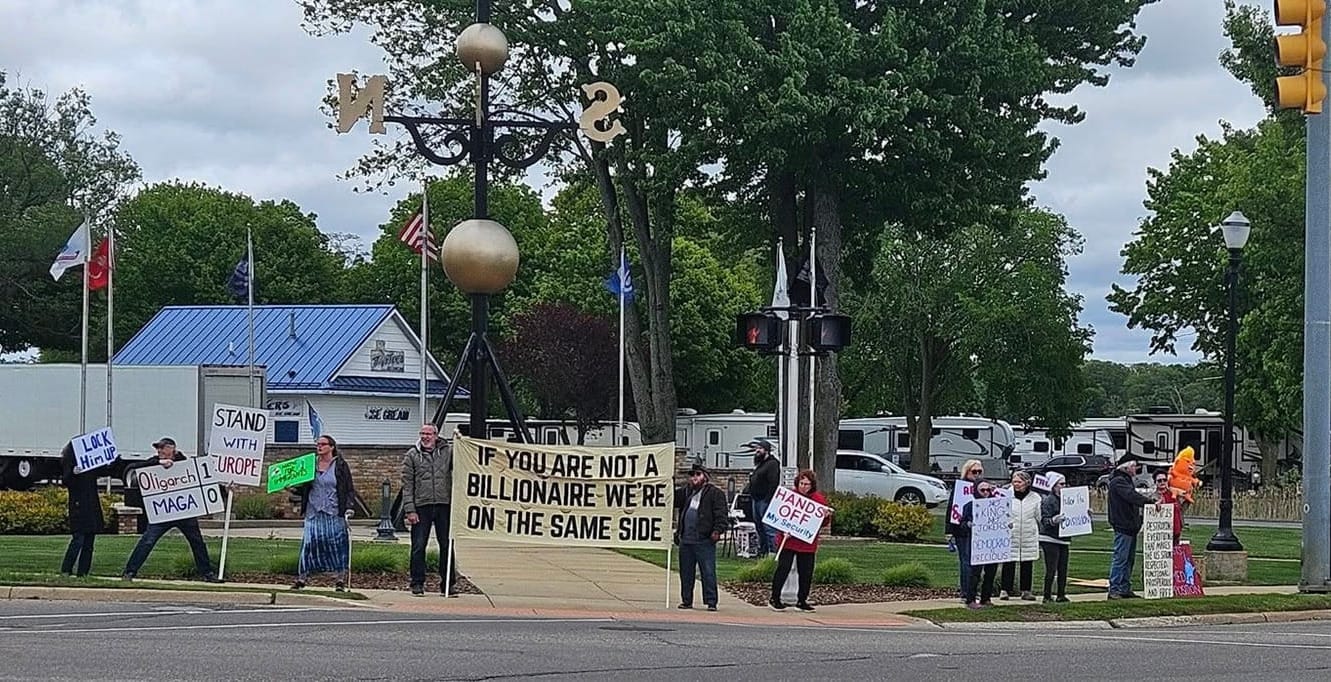 A picture of a line of protesters at the weathervane. The new banner is clearly visible and reads "If you are not a billionaire, we're on the same side"