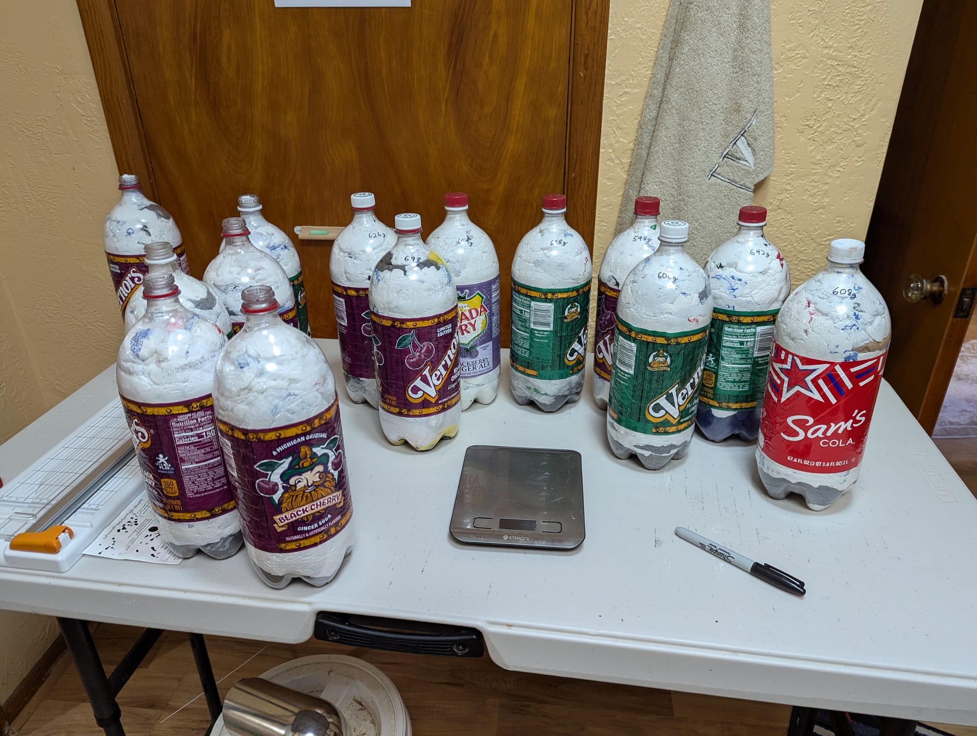 A folding table with 14 complete and partial ecobricks on it. In the foreground is a small kitchen scale, and some of the ecobricks have weights written on them