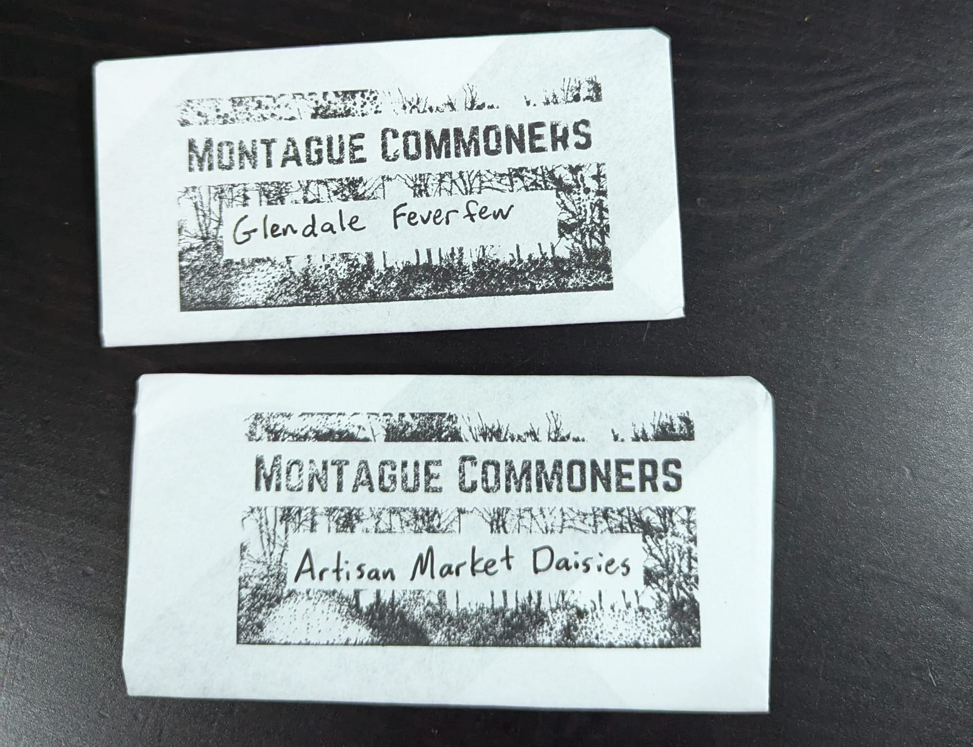 A pair of Montague Commoners seed packets, one labeled "Glendale Feverfew" and one labeled "Artisan Market Daisies"