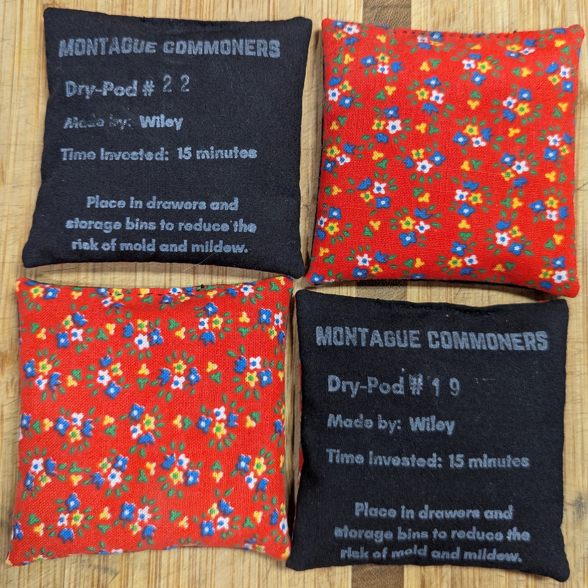 Four square pouches of fabric. Two are pattern-up, and show tiny flowers on a red background. Two are label-up, with information about who made them and how long it took. At the bottom, they say "place in drawers and storage bins to reduce the risk of mold and mildew."
