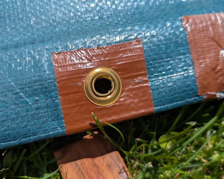 The edge of a green tarp. A section of edge has been reinforced with brown duct tape, and a new brass grommet has been added